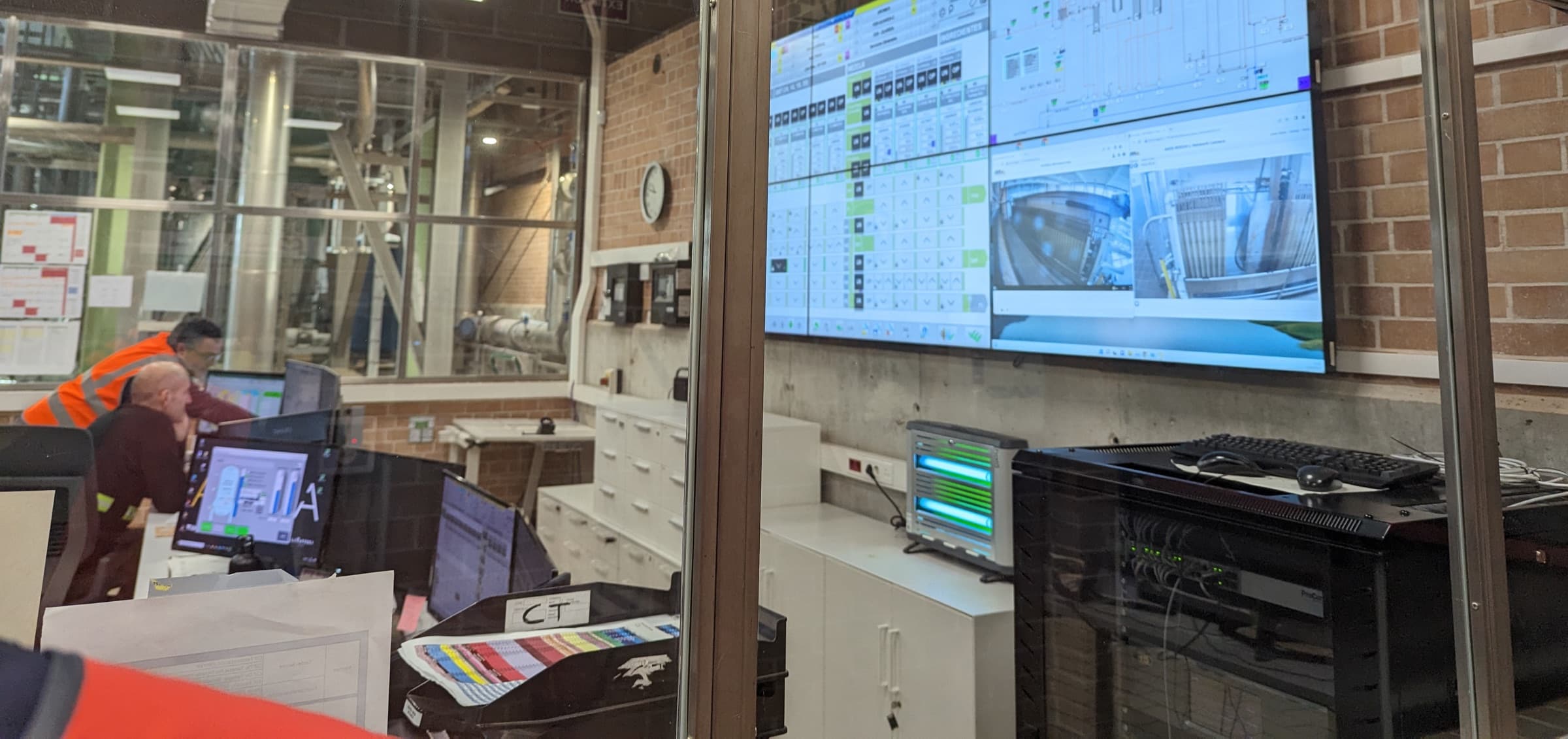 Two operators at a plant control desk, working against a wall of live process displays